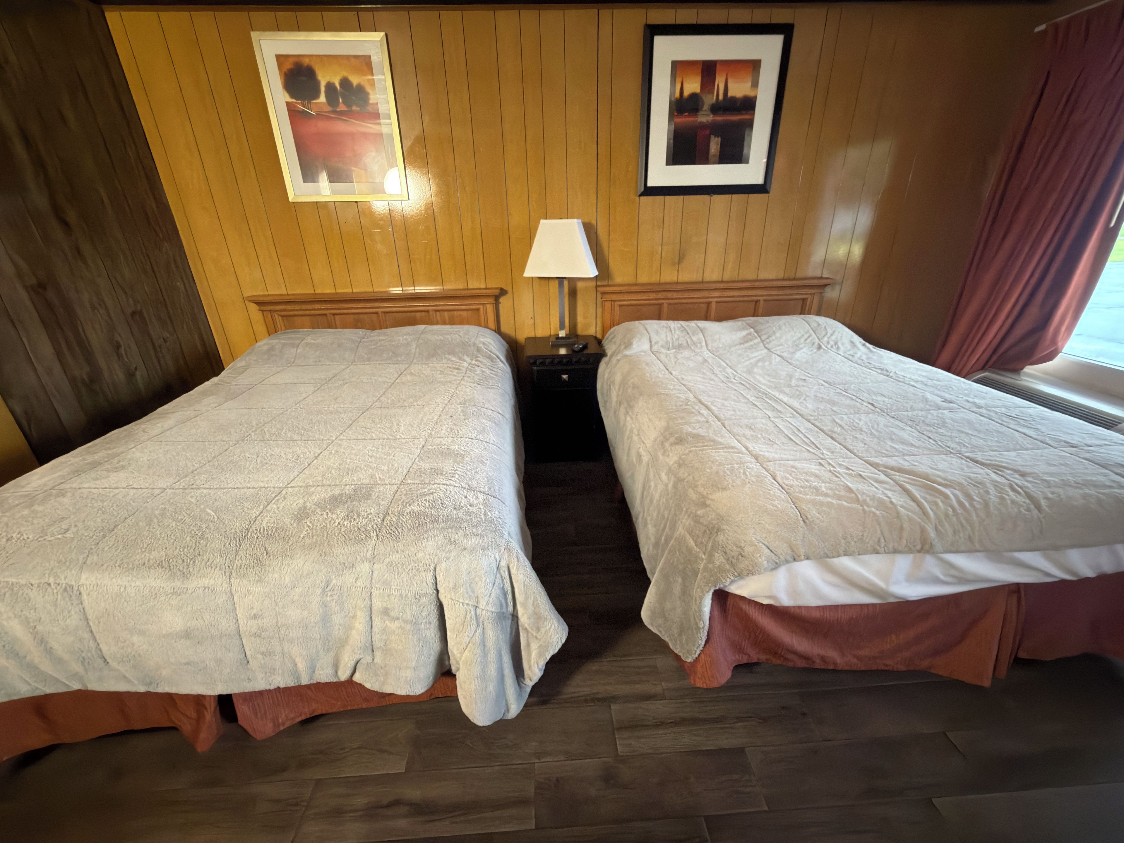 Une chambre d’hôtel avec deux lits, des lambris en bois aux murs, une table de chevet avec une lampe et des tableaux encadrés accrochés au mur.