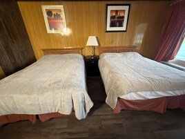 Habitación de hotel con dos camas, paneles de madera en las paredes, una mesita de noche con lámpara y cuadros enmarcados.