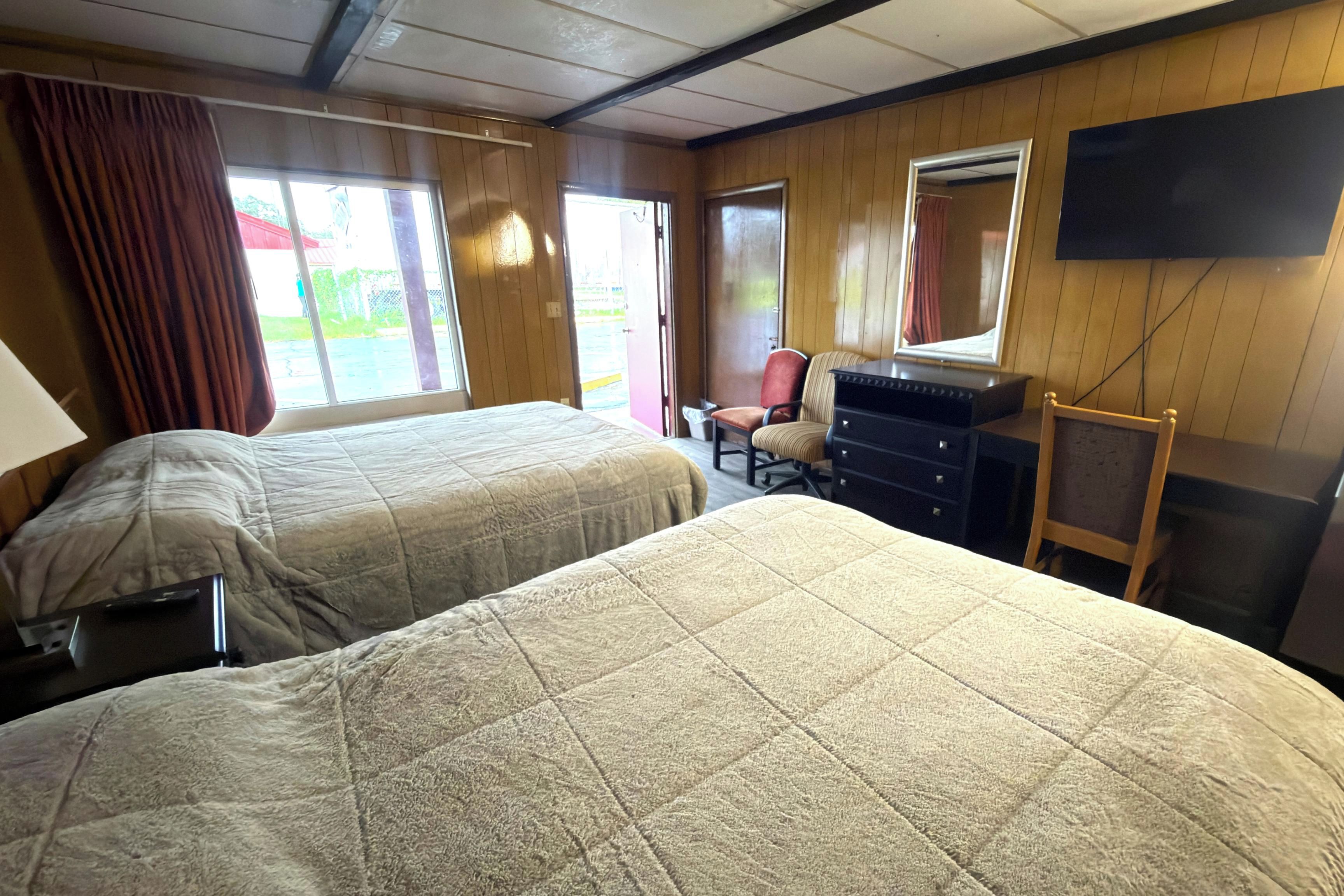 A room with two beds, a desk, a chair, a television, and a window with curtains.