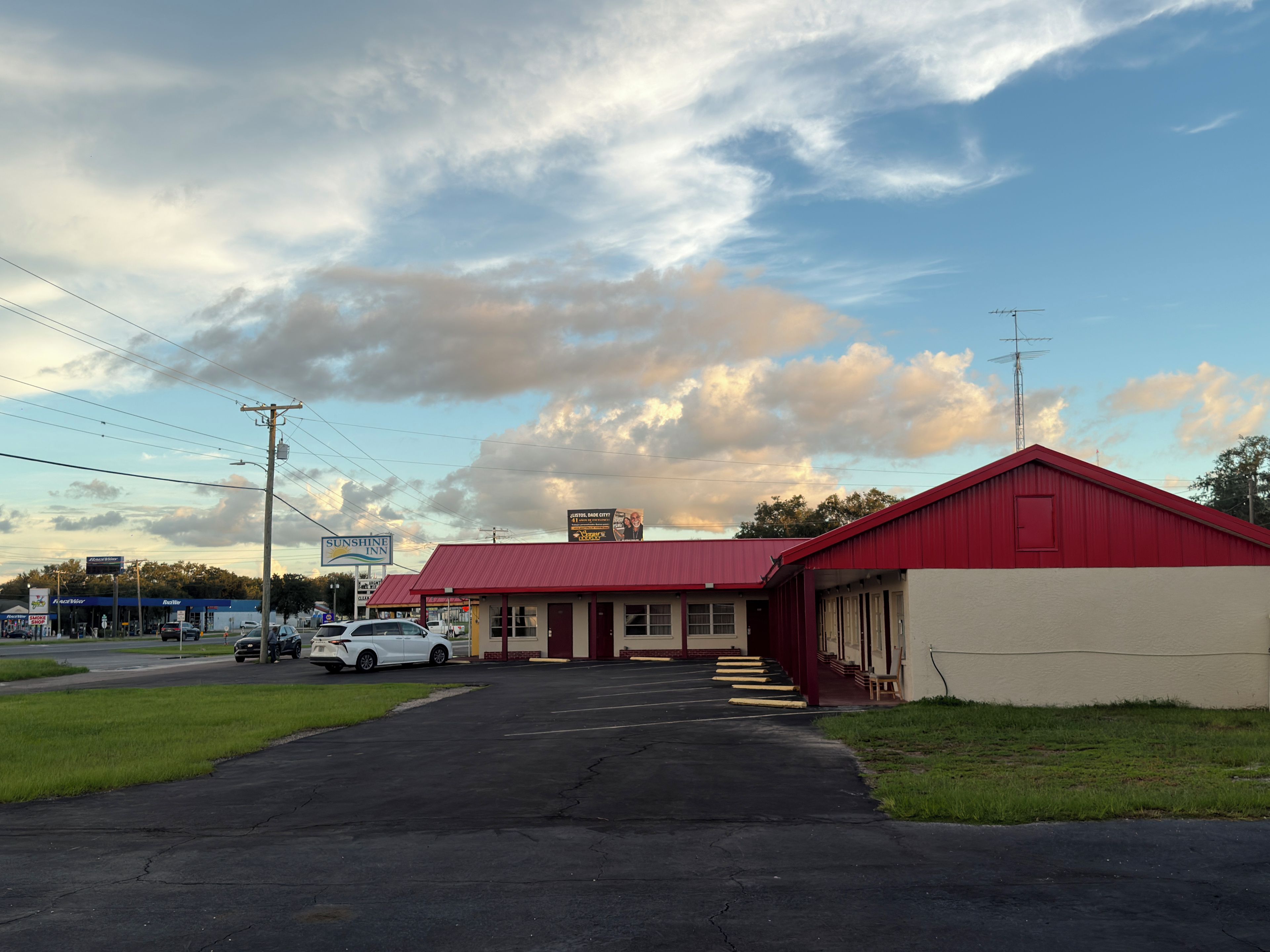 Un motel avec un toit rouge, un parking et un ciel dégagé parsemé de nuages.
