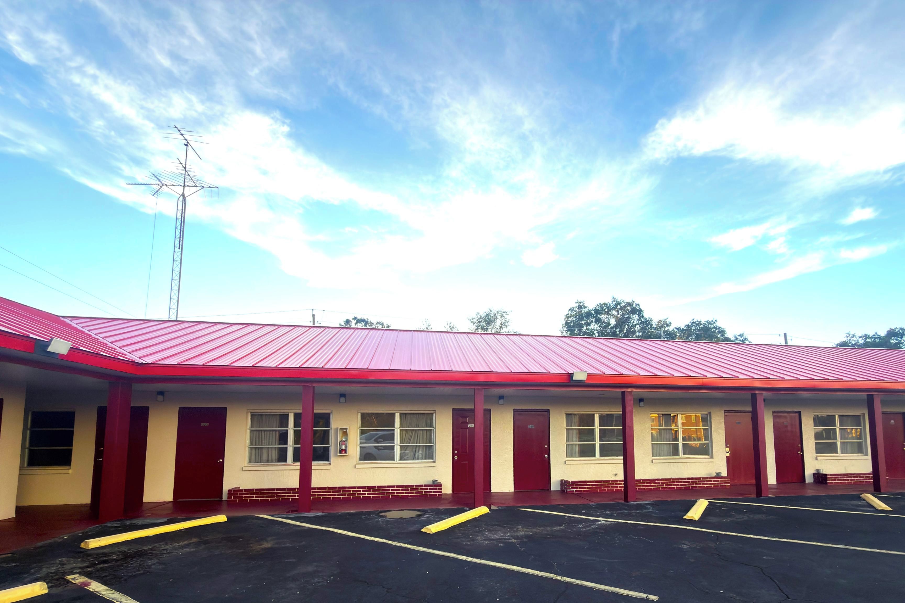 A motel with a red roof and multiple rooms, a parking lot, and a clear sky.