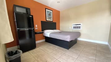 Habitación de hotel compacta con cama, refrigerador negro, basurero y unidad de aire acondicionado mural.