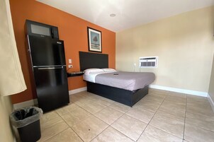 Camera d'albergo compatta con un letto, un frigorifero nero, un cestino e un condizionatore d'aria a parete.