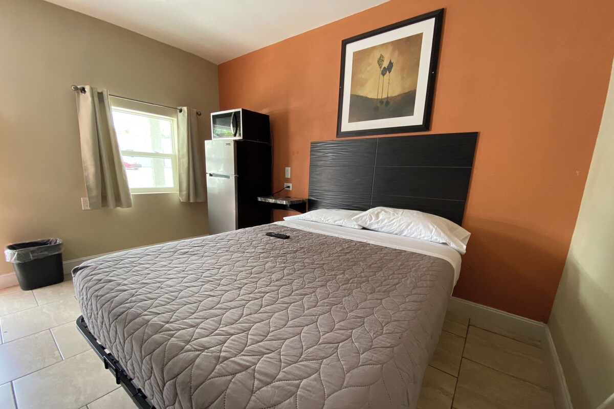 A hotel room with a bed, a nightstand, a trash bin, a window with curtains, and a framed picture on the wall.