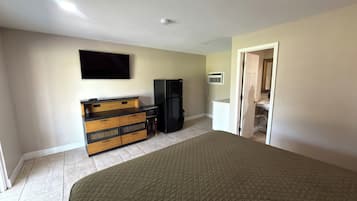 Quarto de hotel com cama, televisão de tela plana, geladeira, cômoda de madeira e porta para um banheiro.