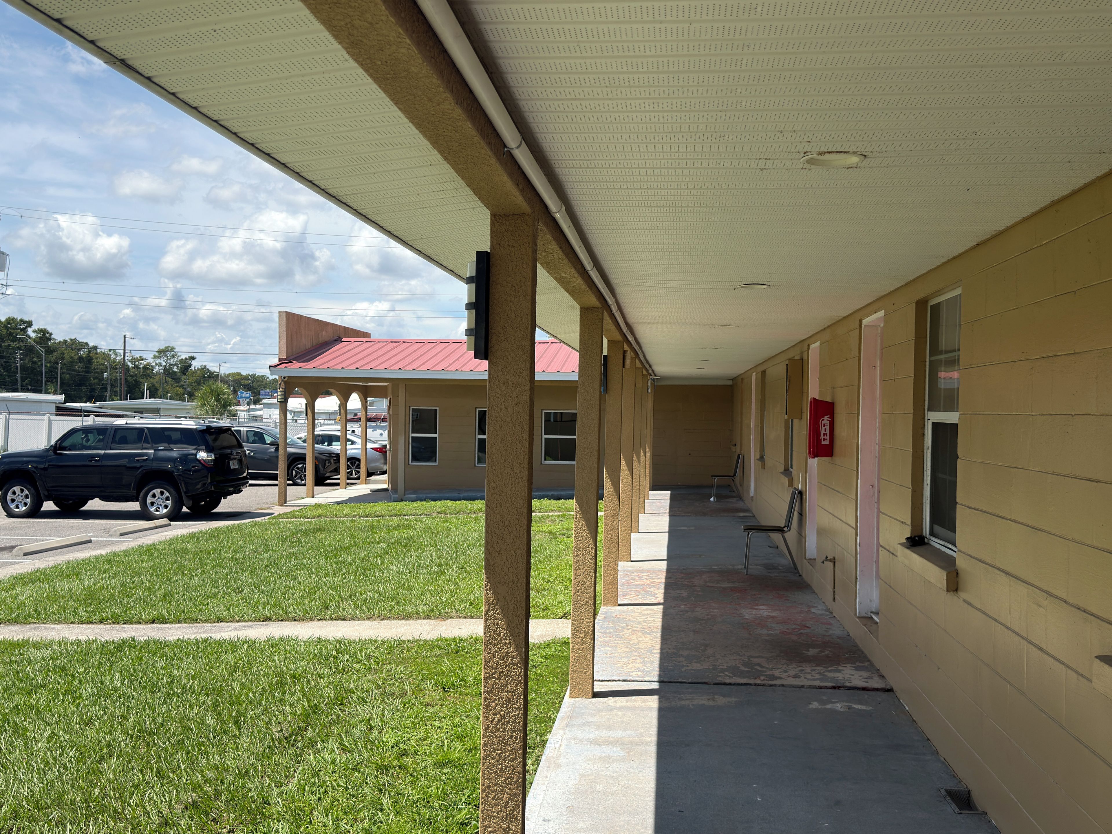 A covered outdoor area with a building in the background, a parking lot, and a grassy area.