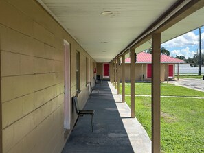 A covered corridor with a row of single chairs, a red door, and a small building with a red roof in the background.
