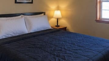 Una cama con un edredón oscuro, dos almohadas blancas y una lámpara de noche sobre una mesita de madera.