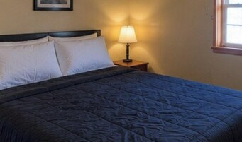 Una cama con un edredón oscuro, dos almohadas blancas y una lámpara de noche sobre una mesita de madera.