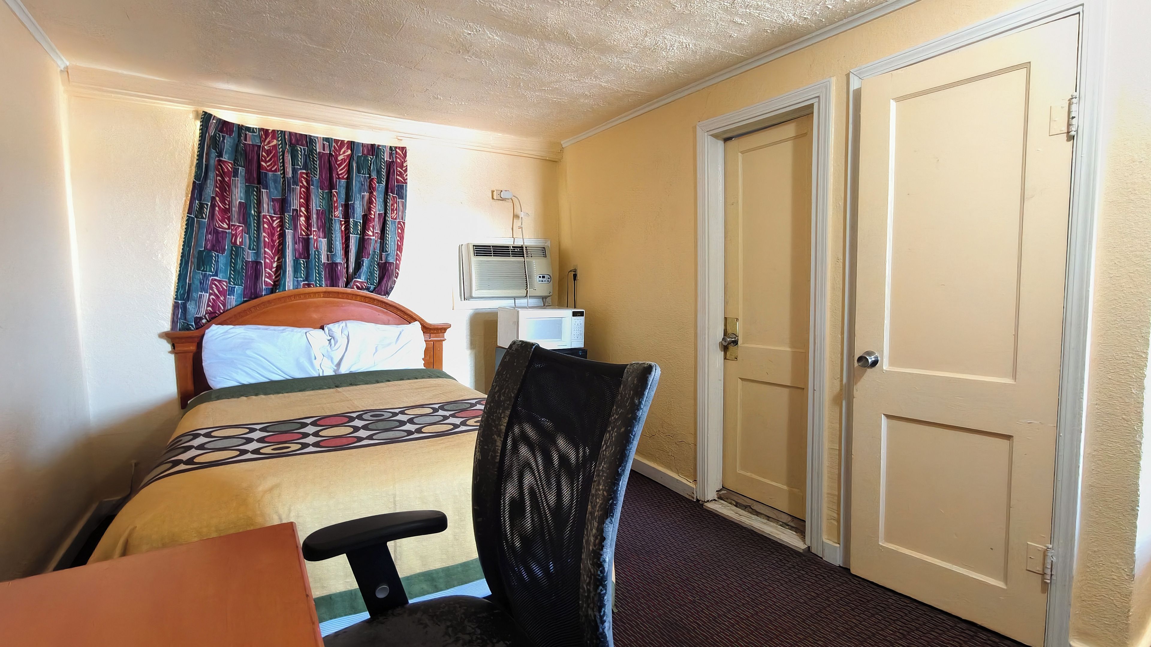 A hotel room with a bed, a chair, a desk, a microwave, and two doors.