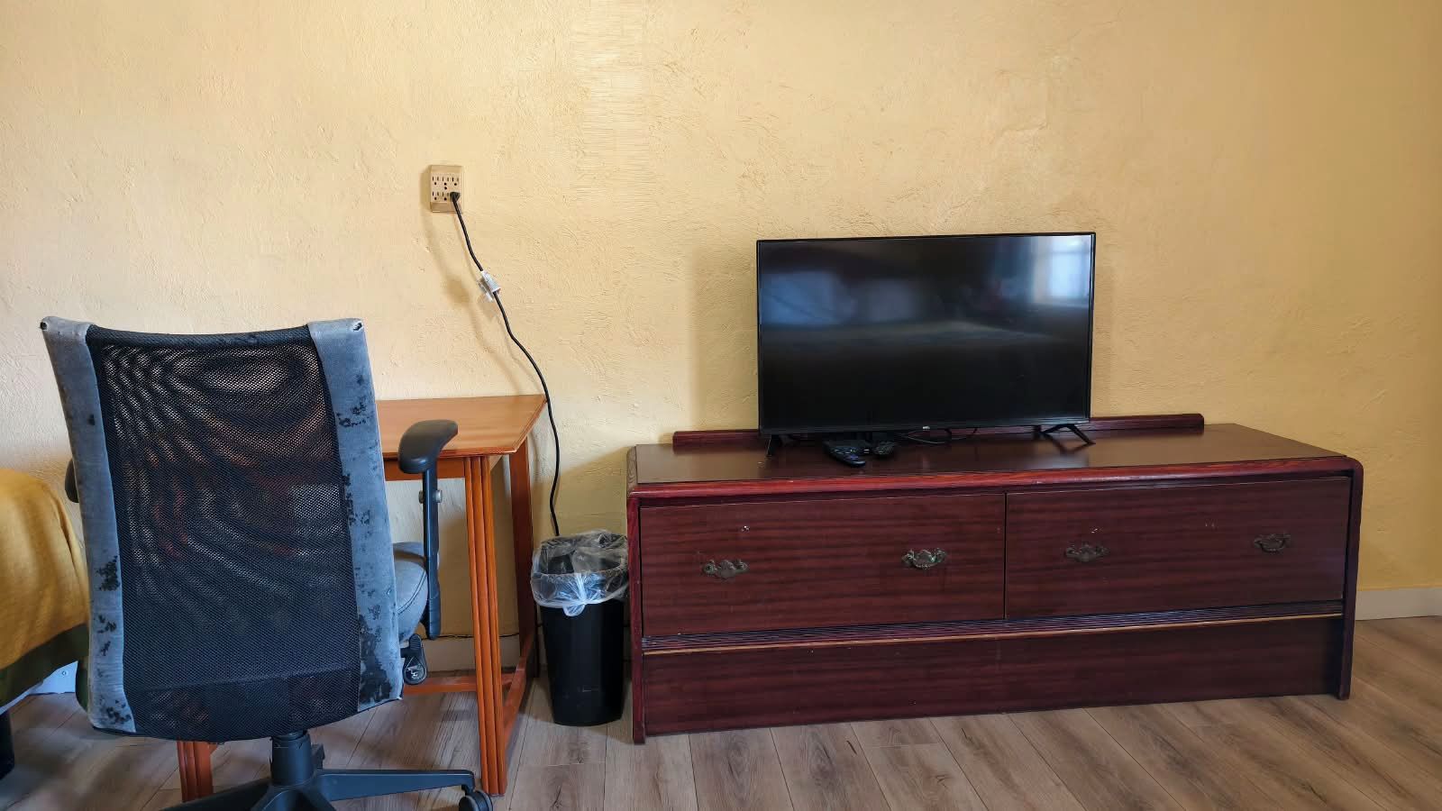 Een kamer met een houten bureau, een stoel, een televisie op een houten dressoir, en een stopcontact.