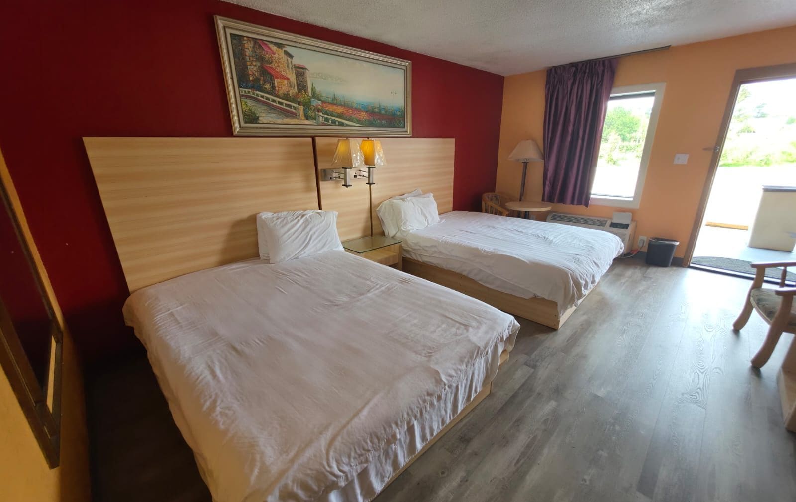 A hotel room with two beds, a painting on the wall, a window with curtains, and a lamp on a bedside table.