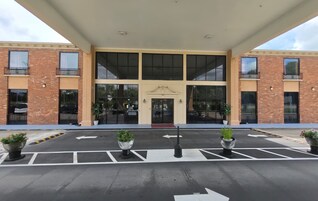 Entrada de un hotel con un corredor cubierto, ventanas amplias y plantas en macetas.