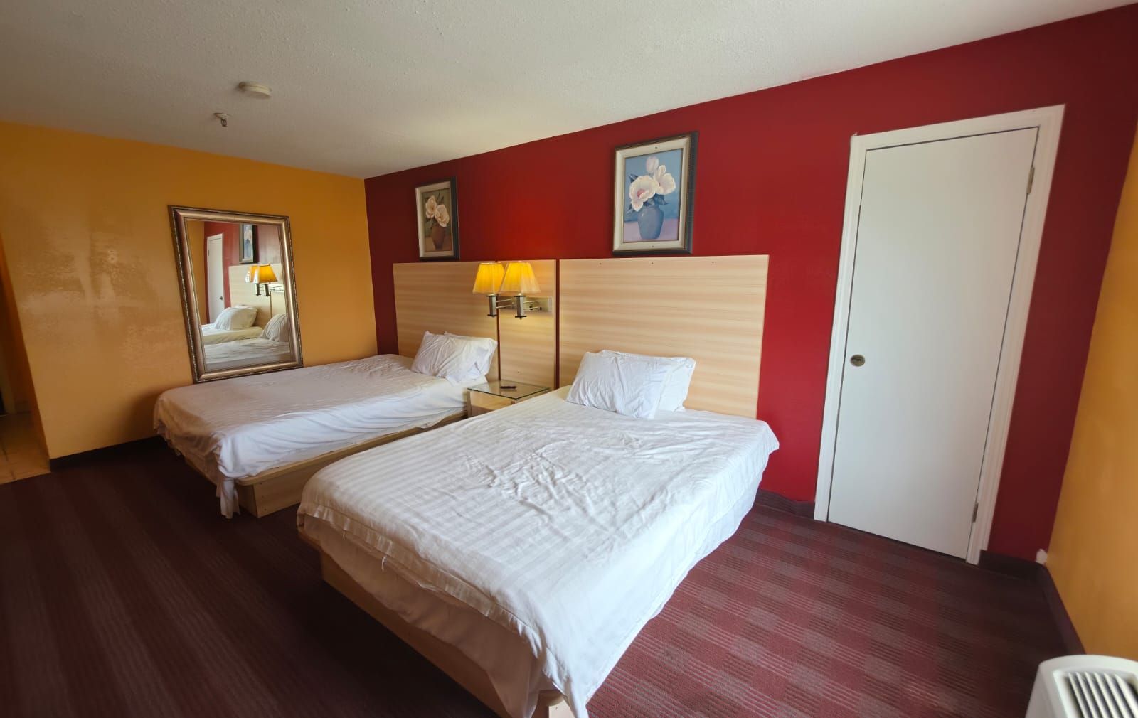 A hotel room with two beds, a mirror, a lamp, and a door.