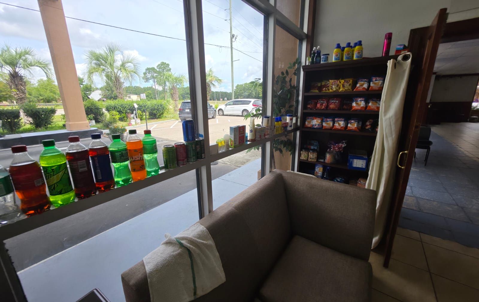 A room with a window overlooking a parking area, a shelf with various snacks, and a chair with a towel.