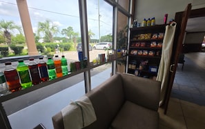 A room with a window overlooking a parking area, a shelf with various snacks, and a chair with a towel.
