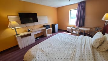A hotel room with a bed, a desk with a flat-screen TV, a chair, a small table, and a window with curtains.