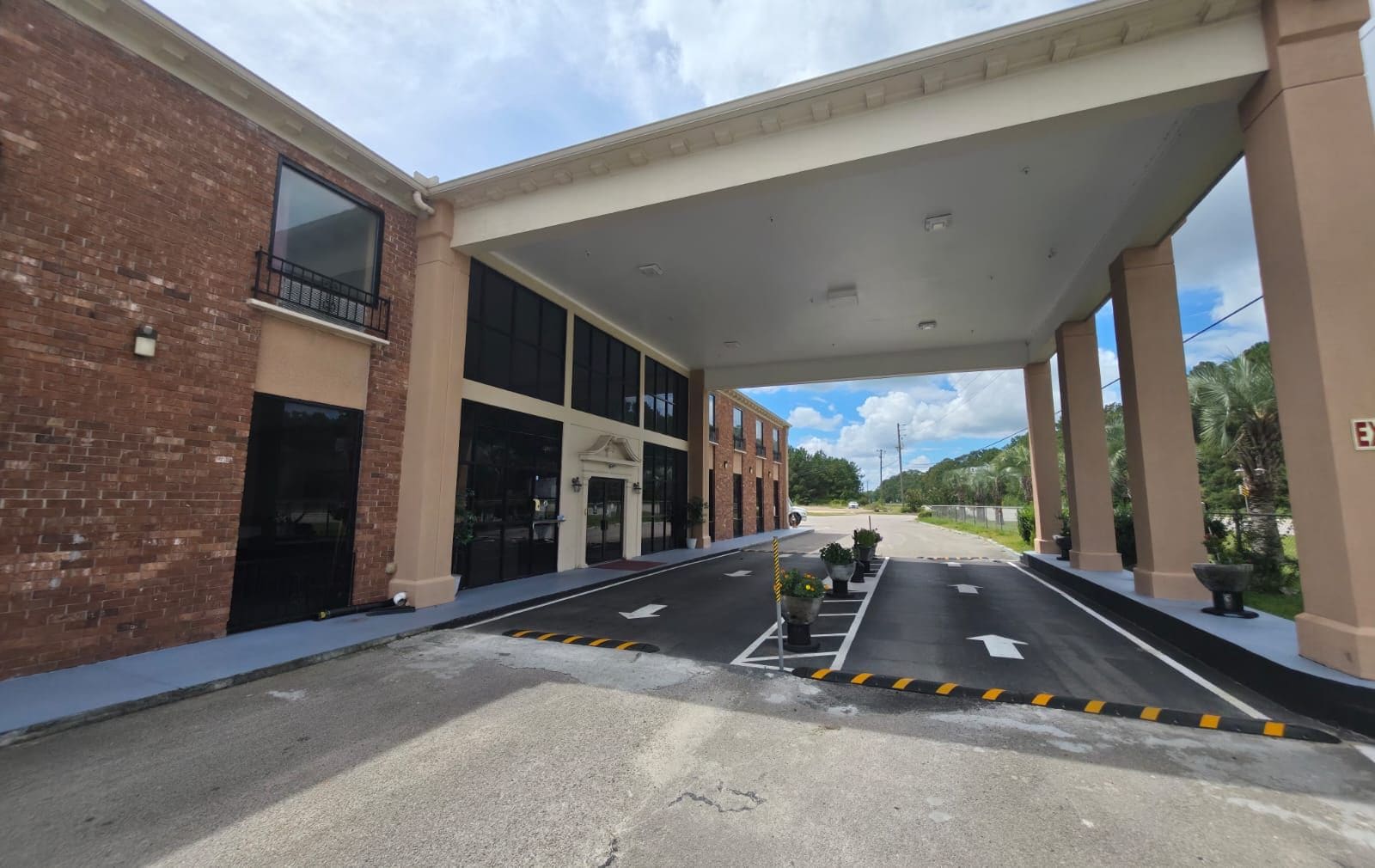 Entrada de un edificio con una marquesina, grandes ventanales y un estacionamiento con flechas direccionales.