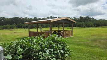 A gazebo with a wooden deck and a grill, surrounded by a grassy field and trees.