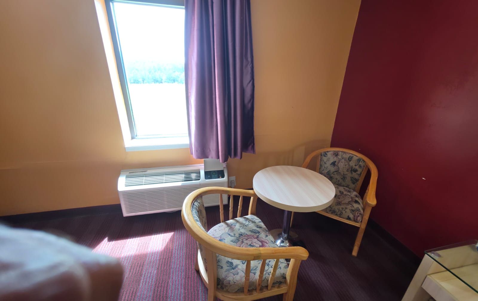 A hotel room with a bed, a round table, two chairs, a window with curtains, and an air conditioning unit.