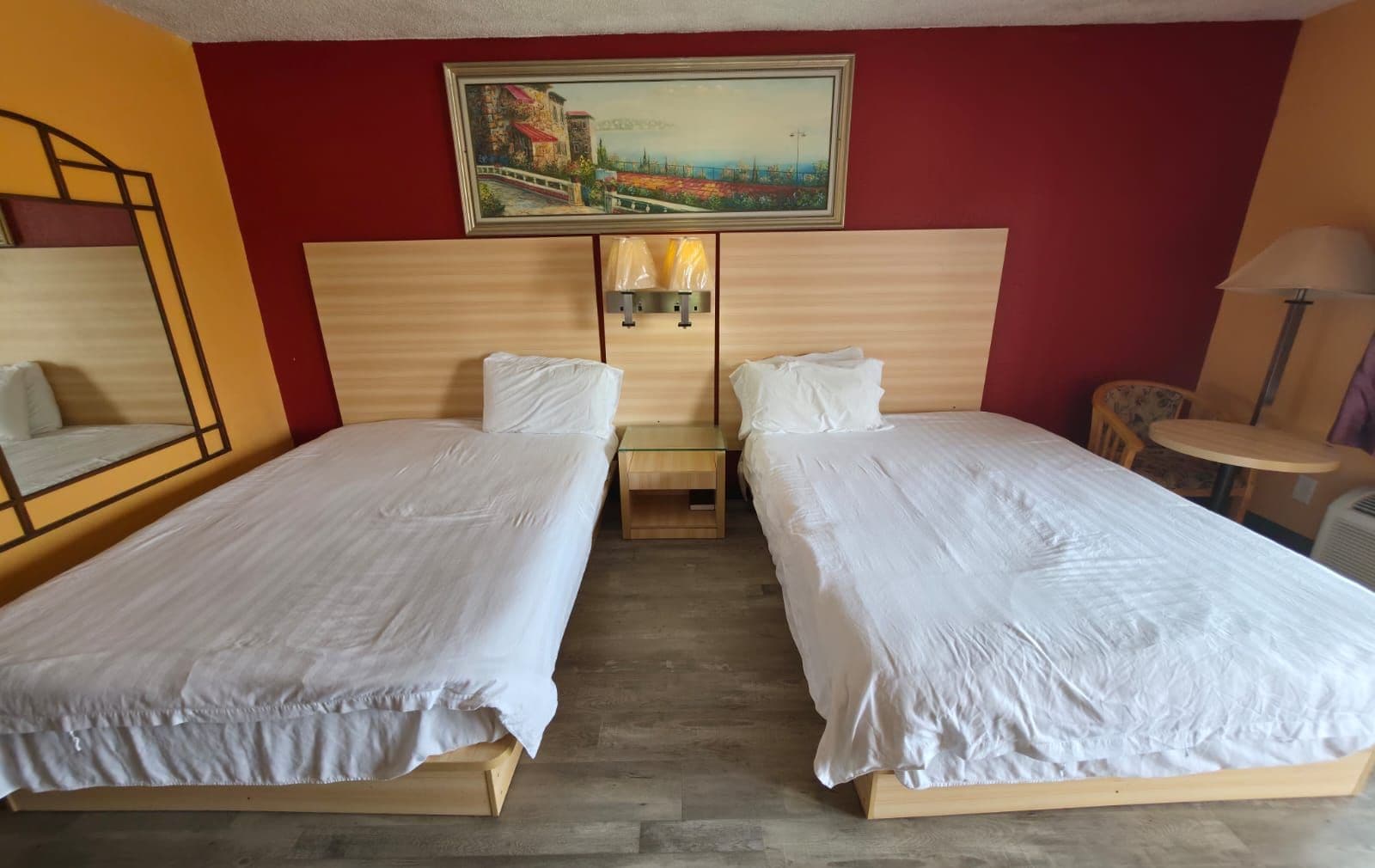 Habitación de hotel con dos camas individuales, cabecero de madera, un cuadro sobre las camas y una pequeña mesa redonda con una silla.