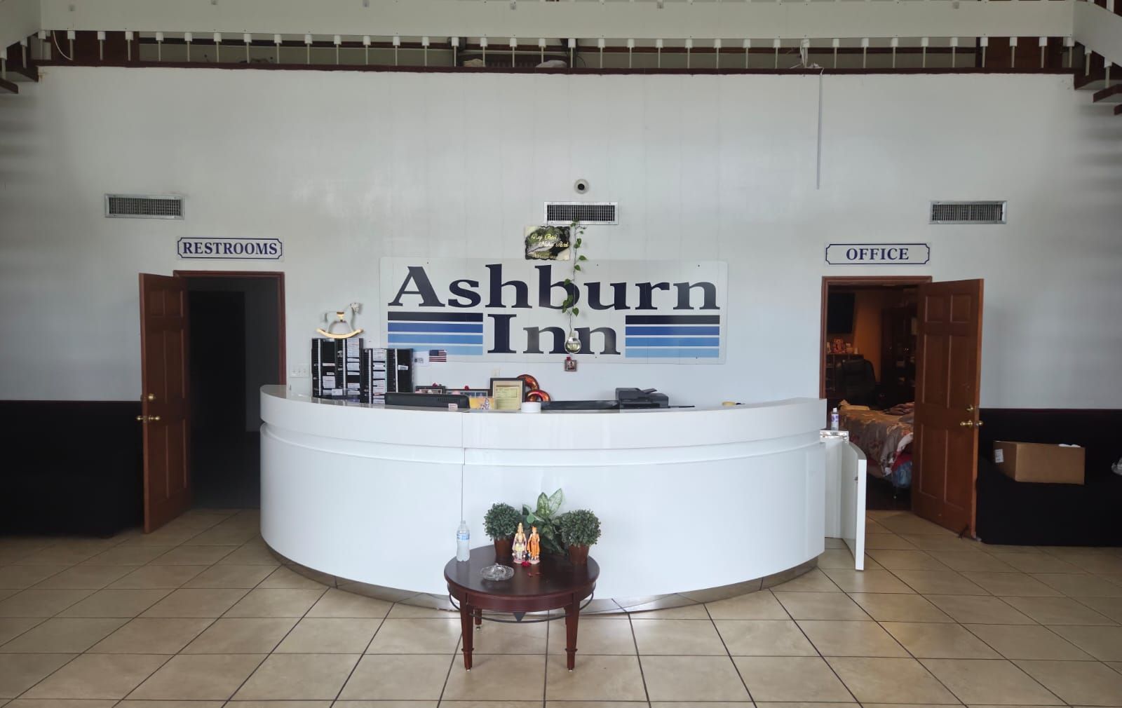 A reception desk at Ashburn Inn with signs for restrooms and office, a small table with a plant, and a tiled floor.