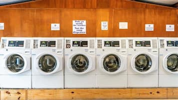 A row of white front-loading washing machines with control panels, arranged in a row against a wooden wall with signs indicating proper use.