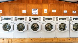 A row of white front-loading washing machines with control panels, arranged in a row against a wooden wall with signs indicating proper use.