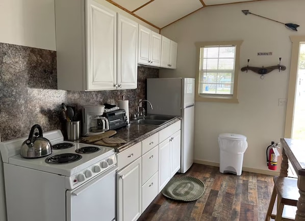 A compact kitchen with white cabinets, a stove, a sink, and a refrigerator. There's a window with blinds and a 'No Smoking' sign on the wall.