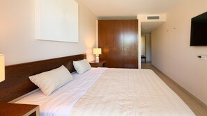 A hotel room with a large bed, a nightstand with a lamp, a wooden wardrobe, and a television mounted on the wall. - Herdade dos Salgados, 3-Bedroom Apartment 8E-1C, Vila das Lagoas (Albufeira)