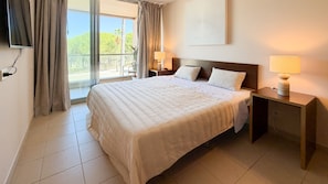 A hotel room with a large bed, a nightstand with a lamp, a view of the outdoors through a sliding door, and a wall-mounted artwork. - Herdade dos Salgados, 3-Bedroom Apartment 8E-1C, Vila das Lagoas (Albufeira)