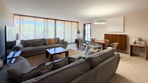A modern living room with a sofa set, a dining table with chairs, and a kitchen area with a wooden countertop. - Herdade dos Salgados, 3-Bedroom Apartment 8E-1C, Vila das Lagoas (Albufeira)