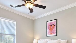 Un dormitorio con cama, ventilador de techo, ventana con persianas, un cuadro colgado en la pared y una mesita de noche con lámpara.