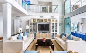 Amplia sala de estar con techo alto, chimenea central y televisor grande. Hay dos sofás blancos con cojines azules, una mesa de centro y una estantería llena de libros y objetos decorativos.