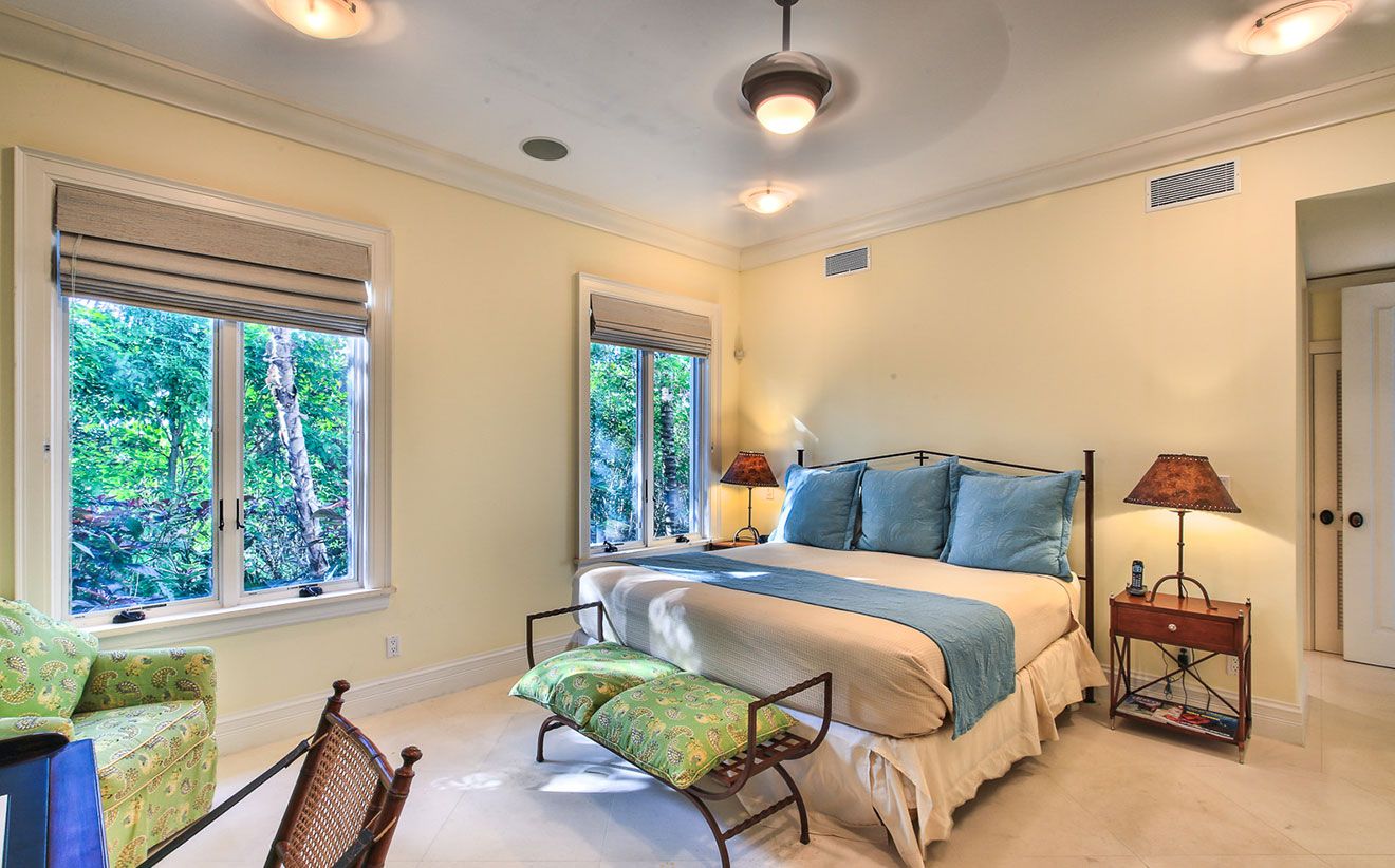 A bedroom with a bed, two windows, a nightstand, a lamp, a chair, and a bench.