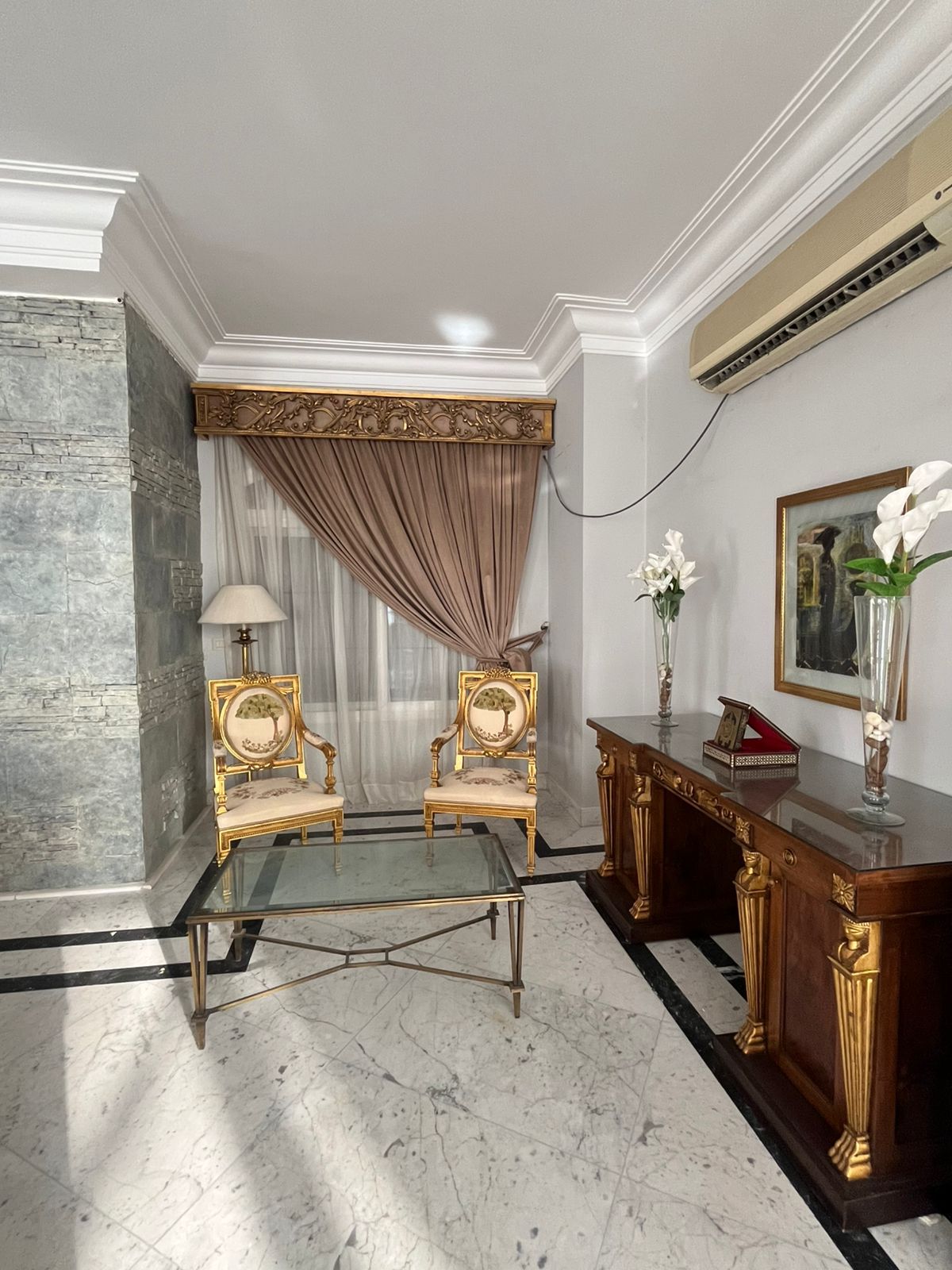 Une pièce comprenant deux fauteuils richement décorés, une table à plateau en verre, un sol en marbre, un mur d’accent en pierre, un rideau drapé, un vase de fleurs et un tableau encadré accroché au mur.