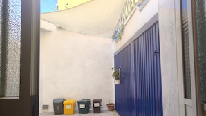 A narrow alleyway with a blue metal gate, a white wall, and a yellow tarp overhead. - Garden, Terrace and Private Parking (Orgosolo)