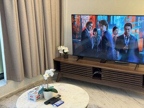 Ein Wohnzimmer mit einem Flachbildfernseher auf einem Holzständer, einem runden Marmortisch mit einer Fernbedienung, einer Taschentuchbox und einem Topf mit einer Orchidee.