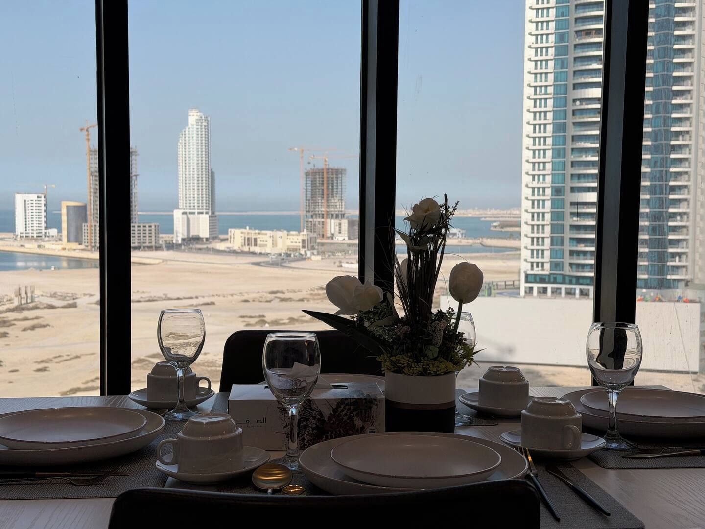A dining table set with plates, glasses, and cutlery, overlooking a cityscape with high-rise buildings and a beach.