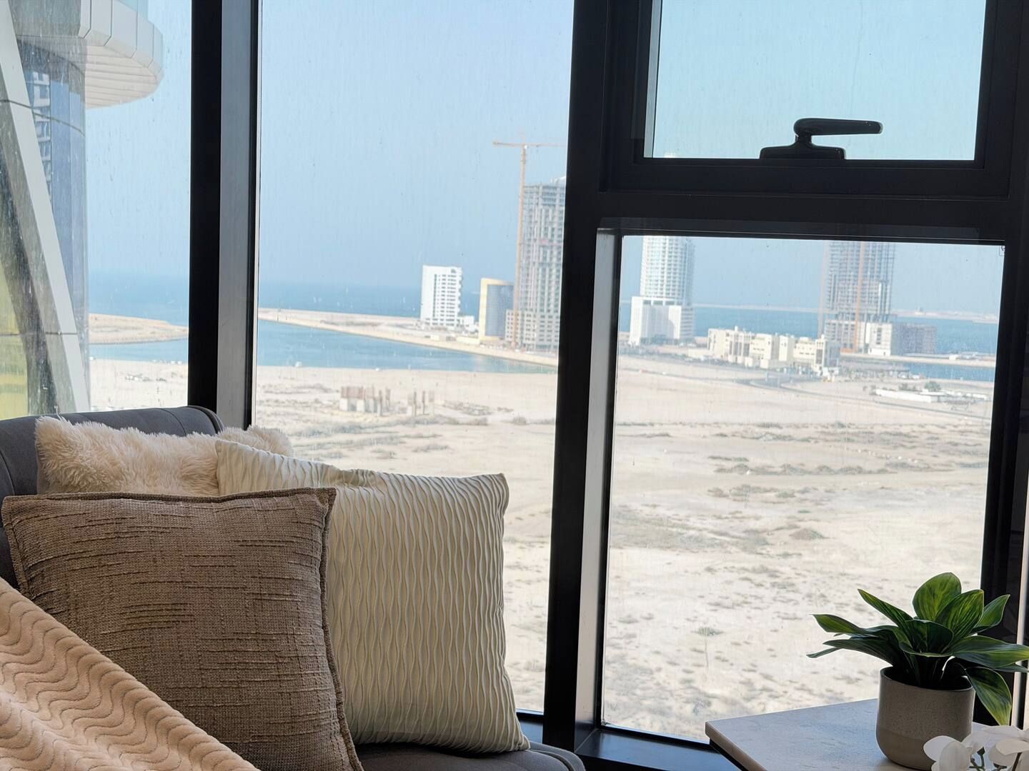 A room with a large window overlooking a beach and cityscape, featuring a sofa with patterned cushions and a potted plant on a side table.