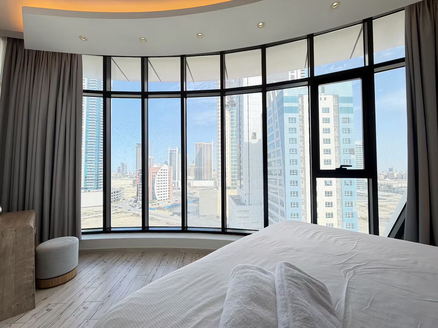 A hotel room with a curved window, a bed, a chair, and a view of a cityscape.