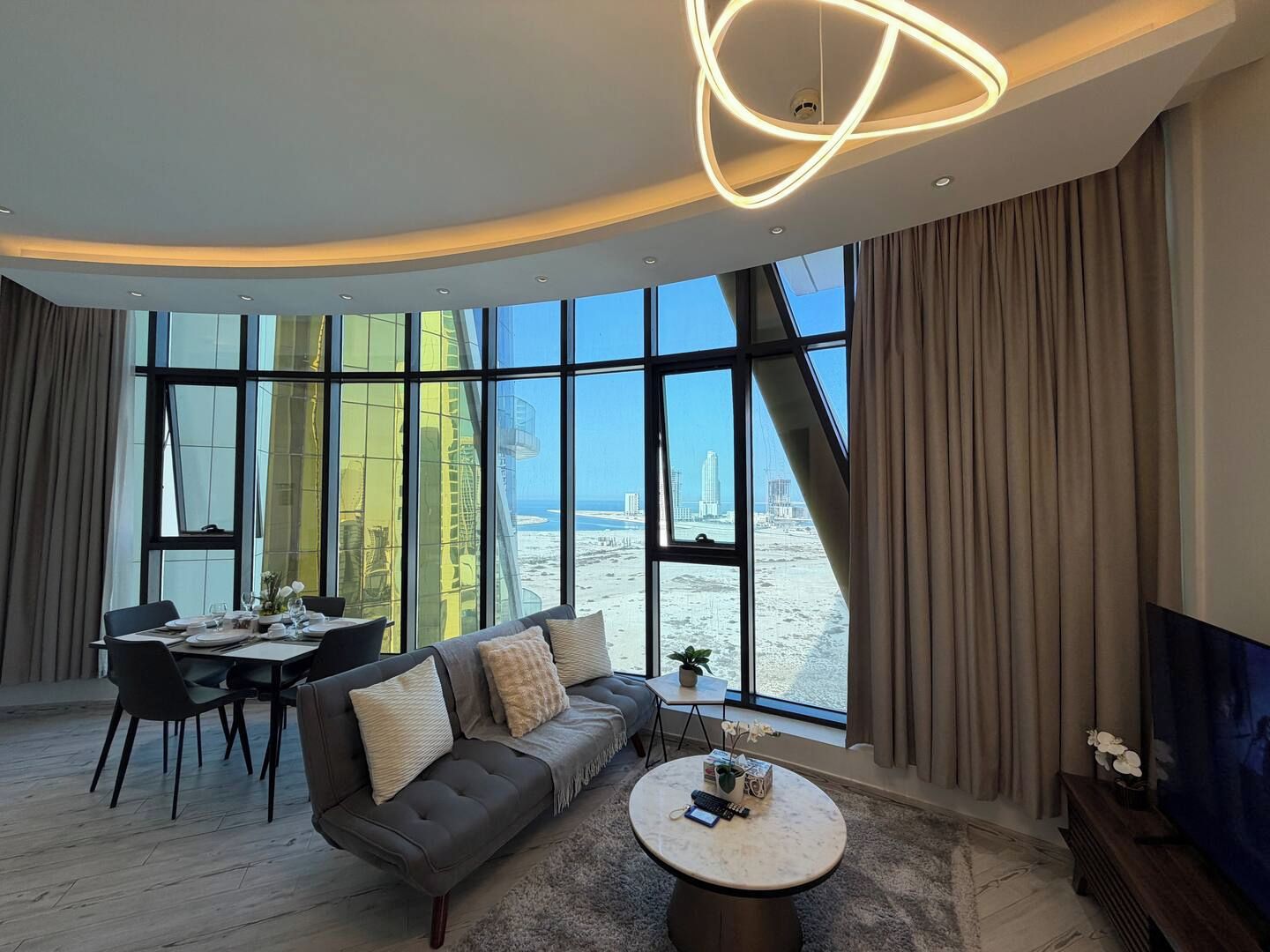 A modern living room with a curved ceiling, a sofa, a dining table, and a view of the beach and city skyline.