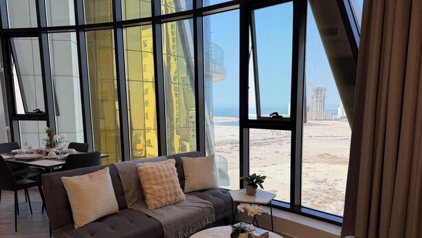 Un soggiorno moderno con divano, tavolino e vista sulla spiaggia e sullo skyline urbano attraverso ampie finestre.
