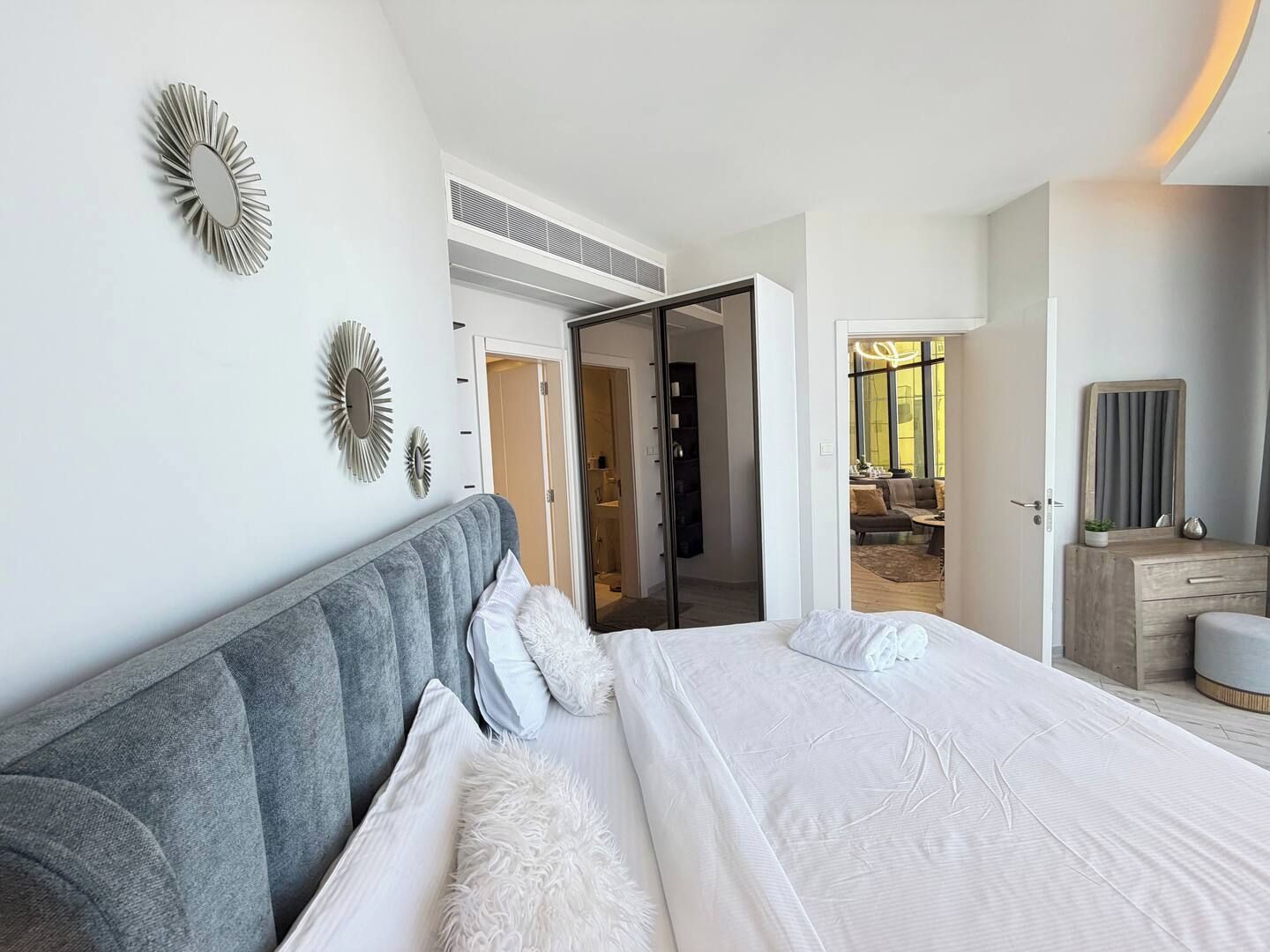 A modern bedroom with a large bed, a grey upholstered headboard, and a view into a dining area.