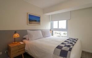 Ein Schlafzimmer mit einem Bett, einem Nachttisch, einer Lampe, einem Fenster mit Jalousien und einem Gemälde mit Strandmotiv an der Wand.