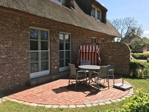 Ein Backsteinhaus mit einem Strohdach, einer Terrasse mit Tisch und Stühlen sowie einem gestreiften Sonnenschirm.