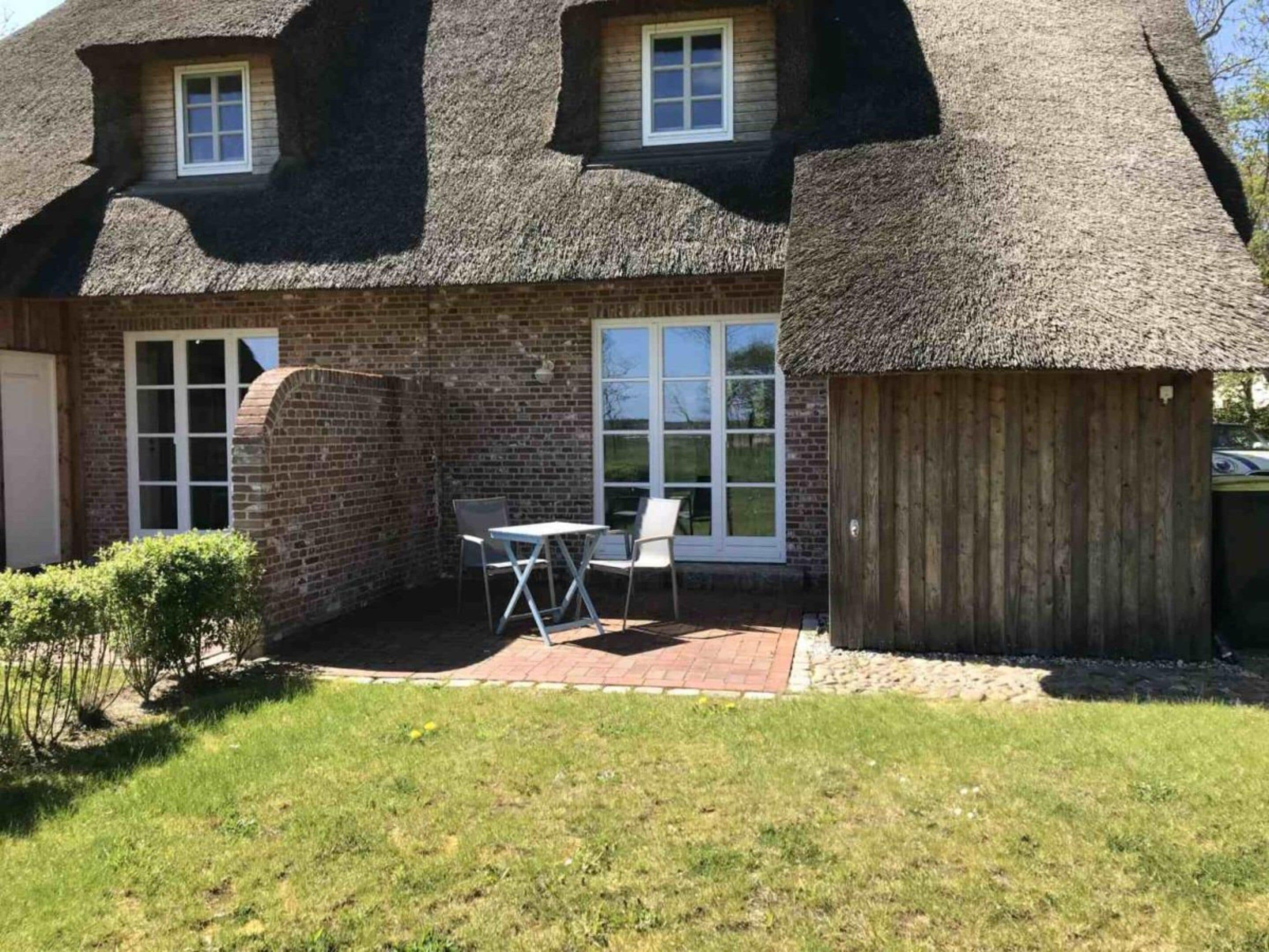 Une maison avec un toit de chaume, des murs en briques et une petite terrasse avec une table et des chaises.
