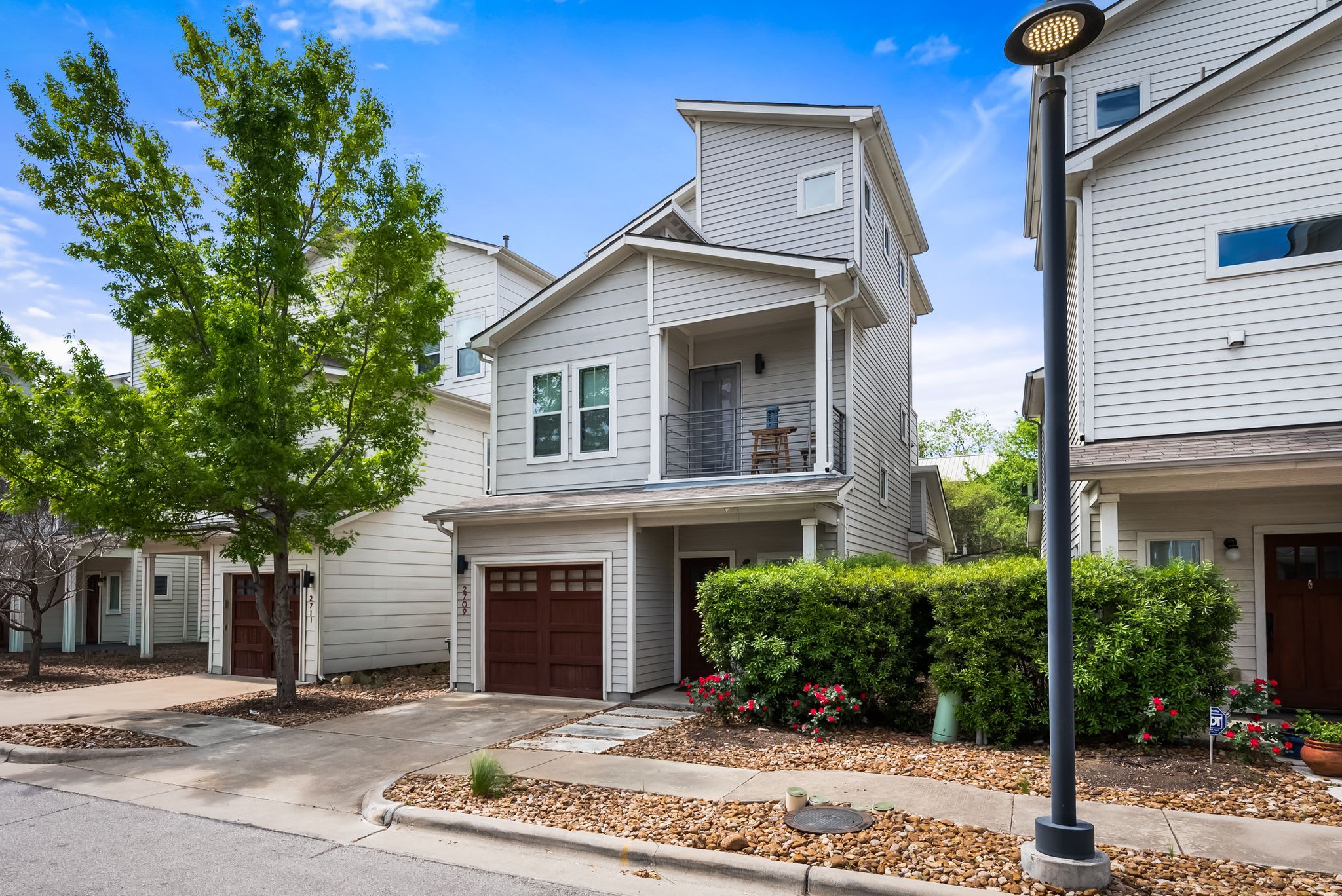 A two-story residential building with a front garage, a balcony, and a well-maintained front yard with shrubs and flowers.