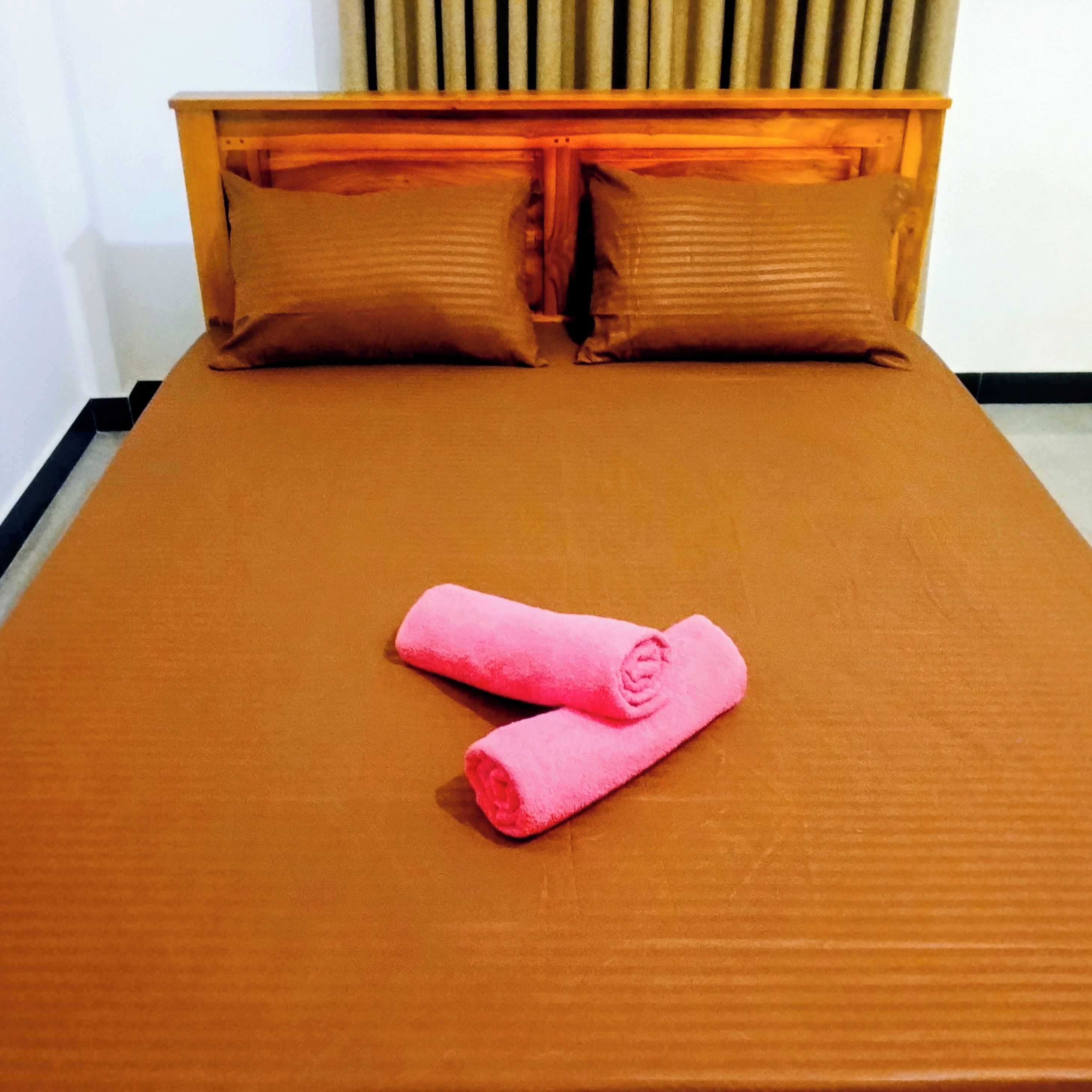 Ein Bett mit einer braunen Matratze und zwei Kissen, ein Holz-Kopfbrett sowie zwei rosafarbene Handtücher.