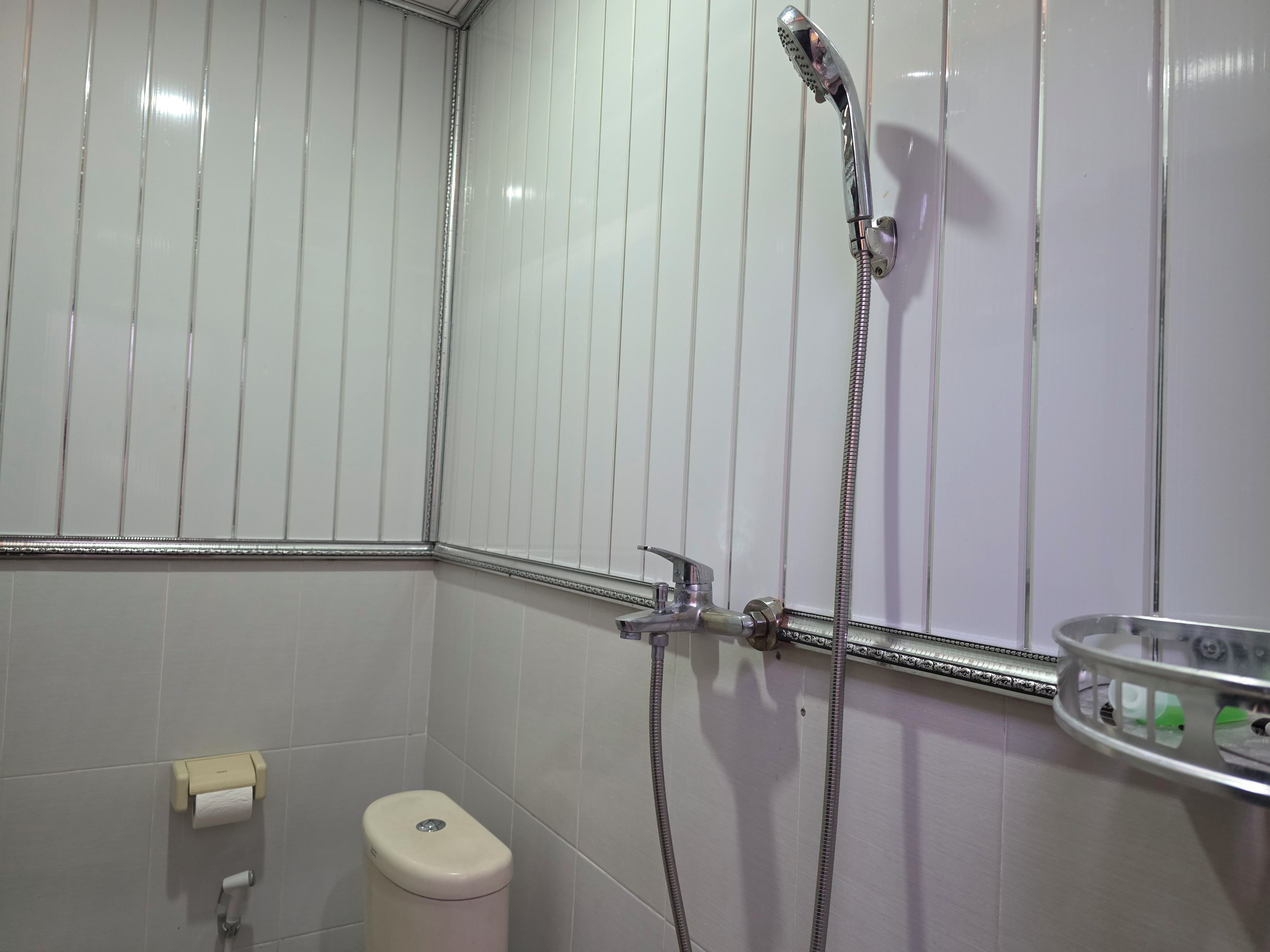 Un baño con inodoro, dispensador de toallas de papel, ducha con cabezal de ducha manual y un recipiente de vidrio en la pared.